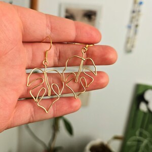 May include: Gold-colored wire earrings in the shape of monstera leaves. The earrings have a hook closure and are held in a hand. The earrings are a minimalist design and are likely handmade.