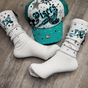 Puede incluir: Una gorra de camionero turquesa con una malla blanca en la parte posterior, que presenta un diseño de tatuaje en blanco y negro con la palabra "Pisces" y pedrería. Dos calcetines blancos hasta el tobillo con diseños de "Pisces" en blanco y negro y pedrería.
