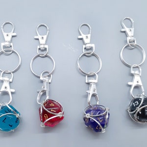 May include: Four keychains with D20 dice. Each die is a different color: teal, red, purple, and black. The dice are encased in a silver wire cage and attached to a silver keychain with a clasp.