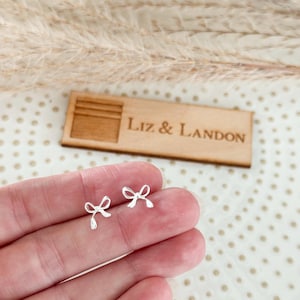 May include: A pair of small, silver-colored bow-shaped earrings. The earrings are held in a hand, with a wooden sign in the background that reads "LIZ & LANDON". The earrings are delicate and detailed.