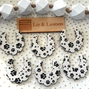 Op de afbeelding: Zes witte, hoefijzervormige houten ornamenten met zwarte pootafdrukken. Een klein houten bord met de tekst "Liz & Landon" is ook aanwezig. Een slinger van witte geometrische kralen is zichtbaar op de achtergrond.