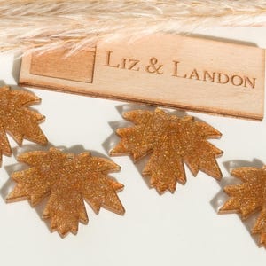 May include: Close-up of several glittery gold maple leaf-shaped ornaments. A wooden sign with the text "Liz & Landon" is visible in the background. The ornaments are on a white surface with dried plants.