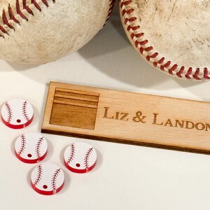 May include: Four white baseball-themed buttons with red stitching and a black baseball seam design. The buttons are on a wooden surface with the text "Liz & Landon" engraved on the surface.