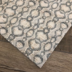 May include: A close-up of a beige and black snakeskin patterned fabric. The fabric has a subtle sheen and a textured surface.