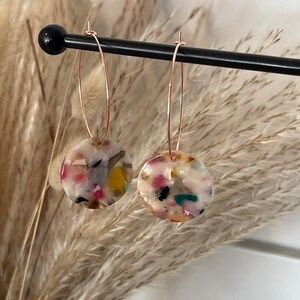 May include: A pair of gold hoop earrings with round, white, multi-colored resin charms. The charms have a speckled, confetti-like appearance.