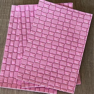 May include: Three pink placemats with a woven texture are stacked on a dark wooden surface. The rectangular placemats feature a repeating pattern of squares with small, evenly spaced holes. The colour is a soft, muted pink.