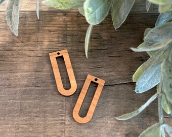 May include: Two unfinished wooden earrings with a rectangular cutout in the centre. The earrings are on a wooden surface.