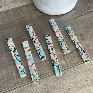 May include: Six colorful, rectangular, marbled acrylic pieces with black swirls. The pieces are arranged in a row on a wood surface.