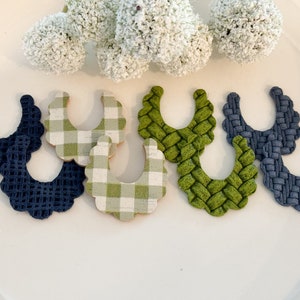 Op de afbeelding: Een verzameling decoratieve slabbetjes in verschillende kleuren en patronen. De items omvatten marineblauw met een geweven textuur, groen en wit geruit en groen met een geweven textuur. Witte bloemaccenten zijn op de achtergrond.