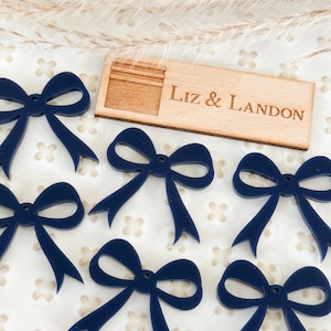 May include: Eight navy blue bow shapes with a hole in the center. The bows are on a white background with a small wooden sign that says "Liz & Landon".