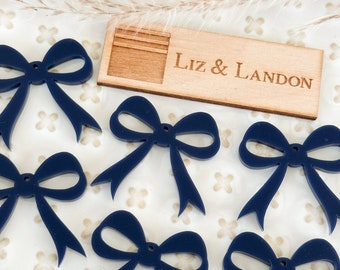 May include: Eight navy blue bow shapes with a hole in the center. The bows are on a white background with a small wooden sign that says "Liz & Landon".