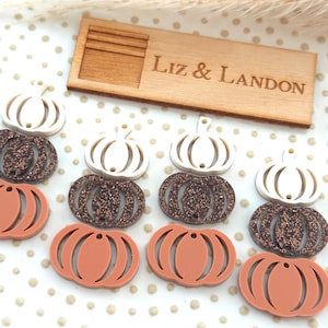 May include: A collection of pumpkin-shaped ornaments in white, brown glitter, and orange. A wooden sign with the text "Liz & Landon" is also present. The ornaments are arranged on a dotted surface, suggesting a fall or harvest theme.