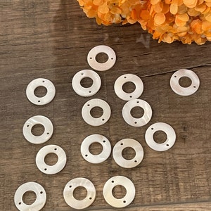 May include: A collection of 15 white, round, shell-like beads with two holes each. The beads are arranged in a semi-circle on a wooden surface.