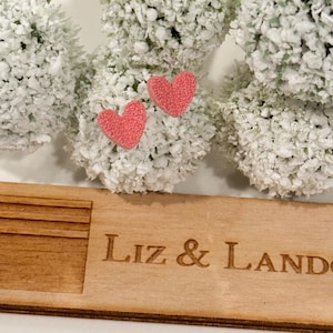 May include: A wooden sign with the text "Liz & Landon" engraved on it. Two small pink felt hearts are placed on top of the sign.