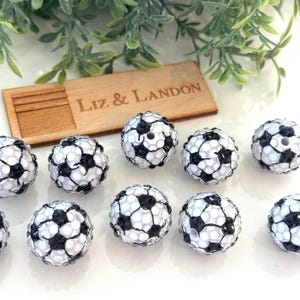 May include: A collection of soccer ball-shaped beads in black and white. The beads are arranged on a white surface, with a wooden plaque that reads "Liz & Landon" in the background. Green foliage is also visible.