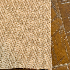 May include: A close-up of a beige woven fabric with a herringbone pattern. The fabric has a textured surface and a subtle sheen.