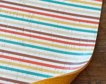 May include: A close-up of a sheet of cork fabric with a striped pattern in shades of brown, red, yellow, and teal. The fabric is folded over to reveal a yellow faux leather backing.