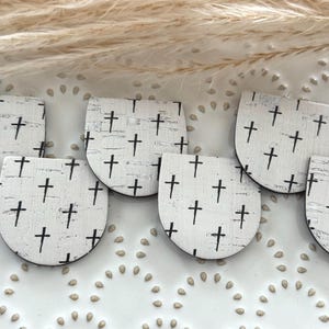 May include: Close-up of several white, semi-circular earrings with a distressed, textured pattern and black crosses. The earrings are arranged on a white surface with a decorative border and dried grass.