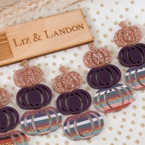 May include: A collection of decorative pumpkin-shaped items in various designs. Some are glittery pink, others are solid dark purple, and some feature colorful striped patterns. A wooden sign with the text "Liz & Landon" is also present.