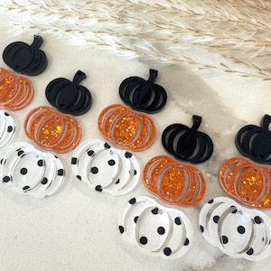 May include: A collection of flat pumpkin-shaped charms made from acrylic. The set includes solid black pumpkins, orange glitter pumpkins, and clear pumpkins featuring black polka dots. Each charm has a small hole at the top for hanging.