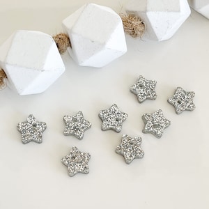 May include: Eight silver glitter star-shaped buttons with a hole in the center. The buttons are scattered on a white surface.