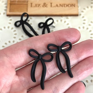 May include: Black bow-shaped earrings. The earrings are matte and have a classic bow design. The image also shows a wooden sign that reads "Liz & Landon".