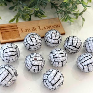 May include: A collection of white and black volleyball-shaped beads. The beads have a textured surface with black lines mimicking a volleyball design. A wooden tag with the text "Liz & Landon" is also visible.