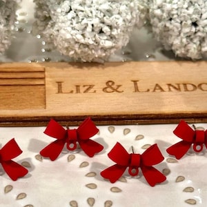 May include: A wooden plaque engraved with "Liz & Landon" sits behind four red bow-shaped earrings. The earrings are on a white surface with a decorative pattern. White floral decorations are in the background.