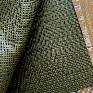May include: Close-up of a textured, olive green material with a woven pattern. The material is partially rolled, revealing its underside. The surface is placed on a wooden table, showing the texture of the wood.