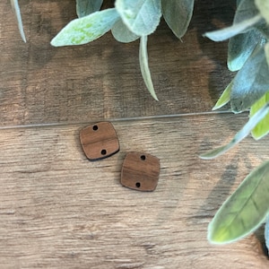 May include: Two small, square wooden beads with rounded corners and two holes each. The beads are a warm brown color and are displayed on a wooden surface, with green foliage in the background. These beads are suitable for jewelry making or craft projects.