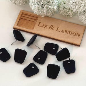 May include: A collection of black stud earrings with silver posts, arranged on a white surface. A wooden box with the engraved text "Liz & Landon" is visible. White floral accents are in the background.