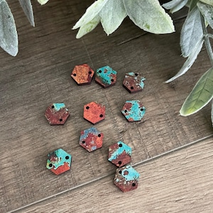 May include: A collection of nine small, flat, hexagonal-shaped buttons featuring a weathered, oxidised metal appearance with a mix of copper-brown and teal-green patina patterns. Each button has two small holes for attachment, arranged in a horizontal line.