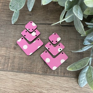 May include: A pair of pink and white polka dot earrings with a geometric shape. The earrings are made of wood and have a layered design.