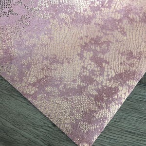 May include: A sheet of material featuring a textured, snakeskin-like pattern in a metallic rose gold finish. The surface displays a mottled appearance with alternating patches of matte dusty pink and shimmering, reflective rose gold scales.