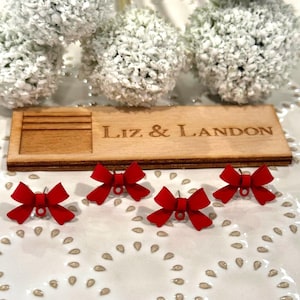 May include: Four red bow-shaped stud earrings with a small loop at the bottom. A wooden sign with the engraved text "Liz & Landon" is also present. The earrings are on a white, patterned surface.