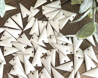May include: A collection of numerous small, off-white triangular charms, each featuring a smooth, glossy finish and a single, centered hole near the top point, arranged in a scattered pile on a textured, light brown wooden surface.