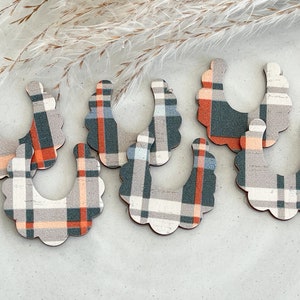 May include: Seven U-shaped wooden cutouts with a plaid pattern in shades of gray, orange, and teal. The edges of the cutouts have a scalloped design. The cutouts are arranged on a light-colored surface, with dried plants in the background.