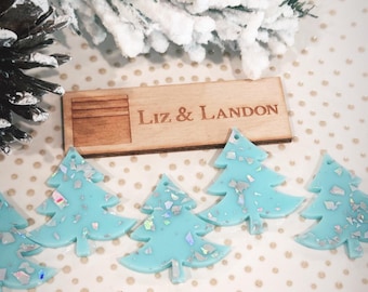May include: Five light blue Christmas tree ornaments with iridescent glitter. The ornaments are on a white background with a wooden sign that reads "Liz & Landon".