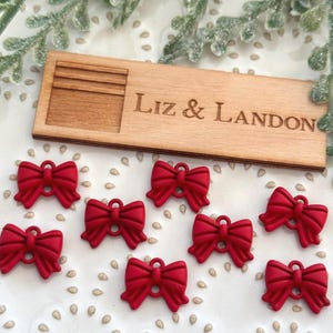May include: A collection of eight small, red bow-shaped embellishments with a loop for attaching. A wooden plaque with the engraved text "Liz & Landon" is also present. The bows are arranged on a white surface.