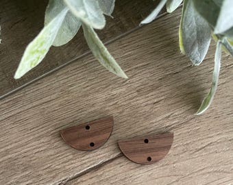 Puede incluir: Dos botones de madera en forma de semicírculo con un patrón de veta de madera marrón oscuro. Cada botón tiene dos pequeños orificios alineados verticalmente en el centro para su fijación. Los botones son planos con bordes lisos y acabado natural.