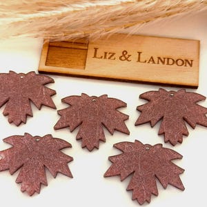 May include: A collection of five reddish-brown maple leaf-shaped wooden ornaments with a textured surface. A small wooden box with the engraved text "Liz & Landon" is visible in the background. Dried ornamental grass is in the background.