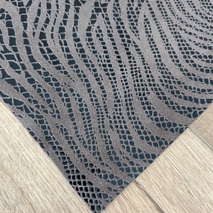 May include: Close-up of a gray, textured fabric with a zebra-like pattern. The fabric has a dark gray backing and a detailed, openwork design. The material is angled across a light wood surface.