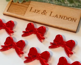 May include: Red acrylic bow embellishments arranged on a white surface. A wooden plaque with the engraved text "Liz & Landon" is visible. Greenery is in the background. These bows are suitable for crafts and decorations.