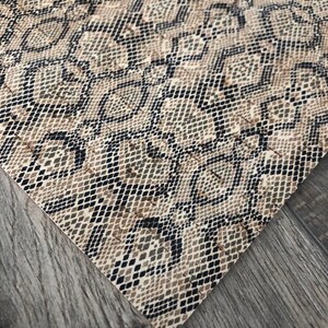 May include: A close-up of a snakeskin pattern fabric. The fabric features a repeating pattern of diamond shapes in shades of brown, tan, and black. The fabric is angled, revealing a portion of a gray wooden surface.