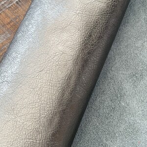 May include: Close-up of a roll of metallic gray leather with a textured surface. The leather is folded over a piece of gray suede. The leather has a subtle sheen and a detailed grain pattern.