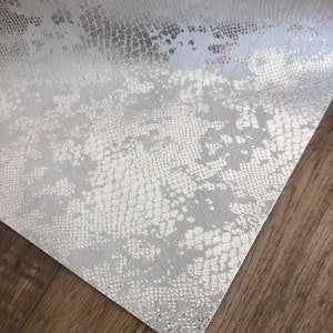 May include: A sheet of silver faux leather with a snakeskin pattern. The material has a textured surface with a metallic sheen, and is angled on a wooden surface.