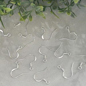 May include: A collection of silver-toned metal earring hooks, each featuring a curved wire design with a small coiled spring detail and a loop at the bottom for attaching jewelry components, scattered across a light gray surface.