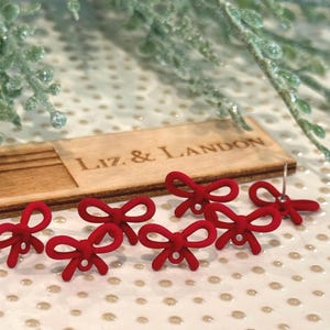 May include: Six red bow-shaped stud earrings with a matte finish are arranged on a patterned surface. A wooden plaque with the text "LIZ & LANDON" is in the background. Decorative greenery is visible in the top of the image.