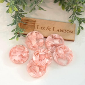 May include: Six pink and white marbled acrylic circle pendants with drilled holes, arranged on a white surface. A wooden box with the engraved text "Liz & Landon" is in the background, along with green foliage.