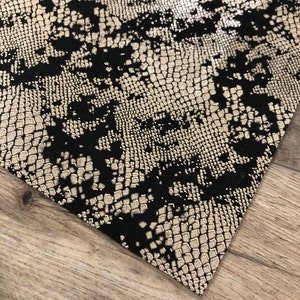 May include: Close-up of a fabric with a snakeskin pattern. The design features a combination of black and metallic gold, creating a textured, reptile-like appearance. The fabric is angled, revealing a glimpse of a wooden surface.